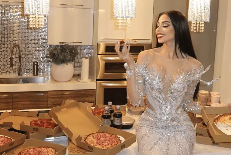Daniela Reyes posando con un vestido de gala de cristales en una cocina mientras sostiene un trozo de pizza.